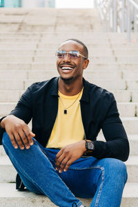 Positive black guy in eyeglasses sitting on white street staircase and looking away happily