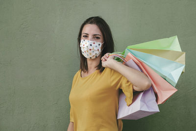 Portrait of woman wearing mask holding shopping bag standing against wall