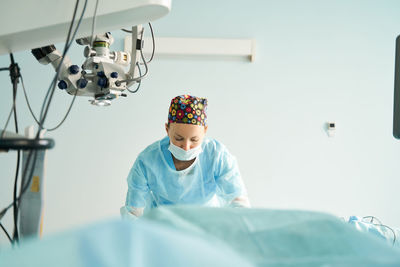 Attentive adult female medic in disposable mask and ornamental cap working in clinic with surgical microscope