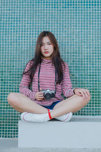 Portrait of young woman sitting with camera against tiled wall