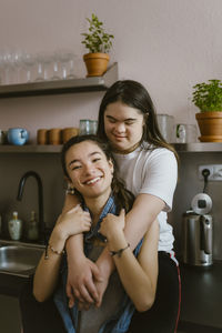 Young woman with down syndrome embracing sister from behind in kitchen at home