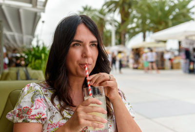 Pretty girl with long hair sitting in bar outdoor in city, drinking lemonade on straw