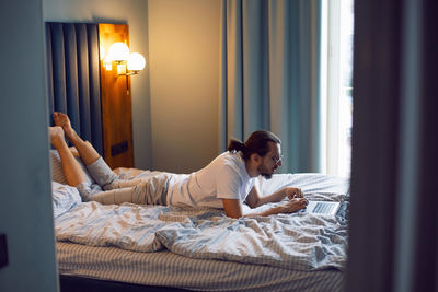 Man in a white t-shirt, a freelancer with glasses, lie on the bed at home with a laptop and works
