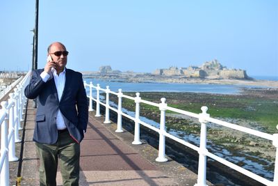 Businessman talking on phone while walking on pier at beach against clear blue sky