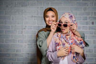 Portrait of smiling woman holding sunglasses on friend face against brick wall