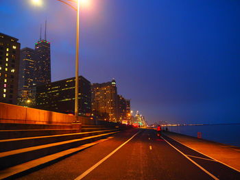 Illuminated road amidst buildings against sky at night