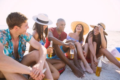 Happy friends holding drinks while sitting at beach against sky