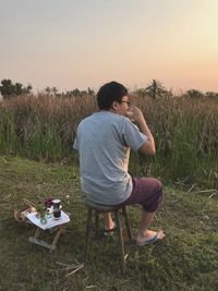 Rear view of man sitting on field against sky during sunset