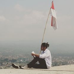 Man reading book while sitting by indian flag against sky in city