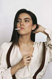 Beautiful young woman smoking cigarette while looking away against white wall