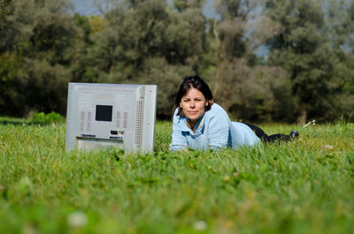 Portrait of woman lying with television set on grass during sunny day