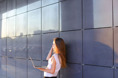 Young businesswoman using laptop standing by wall