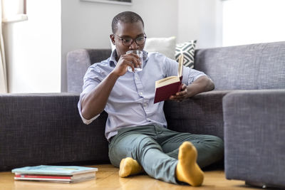 Young man sitting on the floor in the living room reading book and drinking glass of water