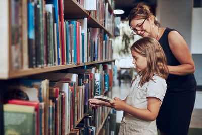 Teacher helping to choose book her schoolgirl in school library. girl selecting books for reading