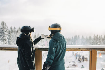 Female friends toasting coffee cups while standing by railing during winter