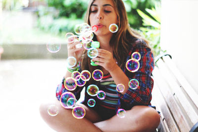 Young woman blowing soap bubbles while sitting on bench