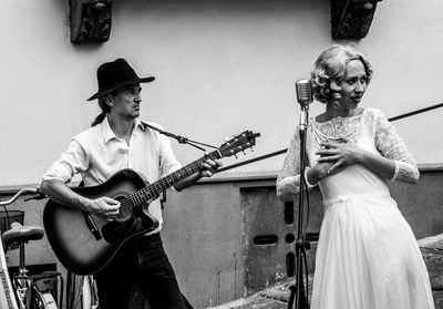 Woman singing on microphone with man playing guitar against wall