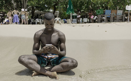 Black traveler sitting on the sand near the sea and browsing social networks on cell phone