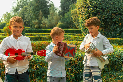 Brothers playing musical instruments while standing against plants in park