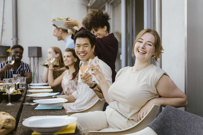 Portrait of happy young woman holding wineglass while sitting with group of friends at dinner party in balcony