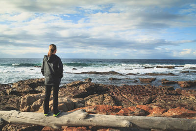 Rear view of woman standing on log by sea against cloudy sky