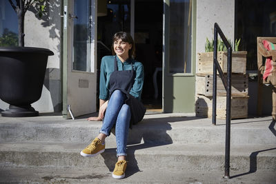 Full length of smiling female owner sitting on steps outside store