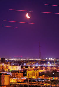 Airplane trails cross the crescent moonlit sky above the urban landscape with a tower on the horizon