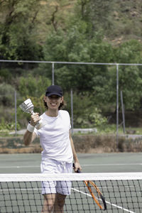 Happy and smiling young amateur tennis player looking to camera winning an award. holding a cup 