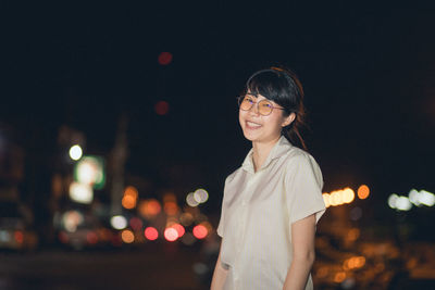 Portrait of smiling young woman standing against sky in city at night