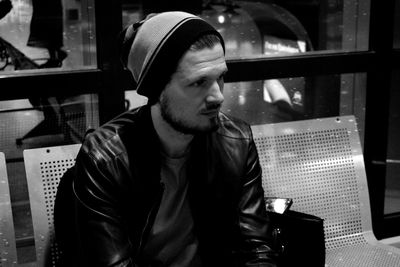 Man wearing knit hat looking away while sitting on bench in city