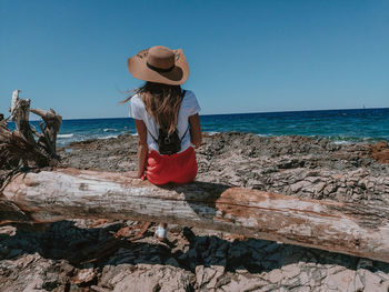 Rear view of woman sitting on driftwood at beach against sky