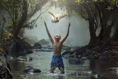 Shirtless boy with arms raised looking at bird flying standing in river