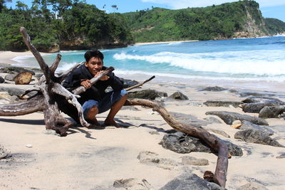 Portrait of young man crouching by driftwood at beach during summer