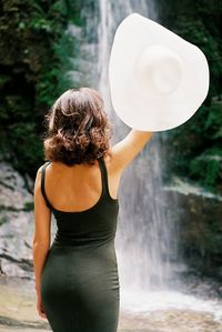 Rear view of woman holding hat with waterfall in background