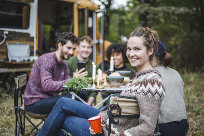 Portrait of smiling blond woman sitting friends at table by caravan in forest during camping