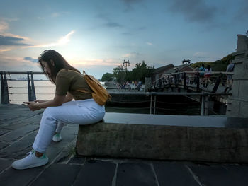 Young woman sitting on railing against sky during sunset