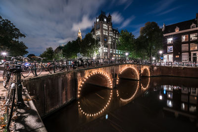 Bicycles parked on illuminated arch bridge over canal against cloudy sky at dusk
