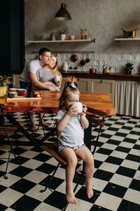 A little girl is holding a mug drinking tea and her parents are kissing from behind in the kitchen 