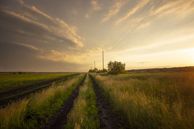 Dirt road amidst field against sky during sunset
