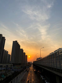 Cars on road by buildings against sky during sunset