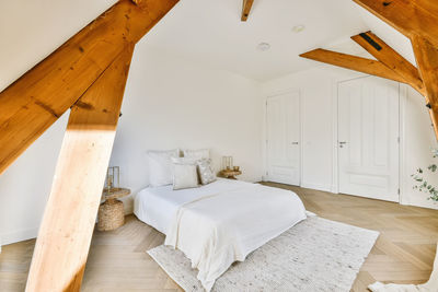 Comfortable bed with soft pillows and creative wicker bedside tables in spacious light bedroom with white walls and wood beams on sunny day