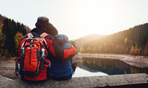 Rear view of couple wearing warm clothing sitting by lake during sunset