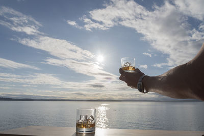 Cropped image of man hand holding whiskey glass by sea on sunny day