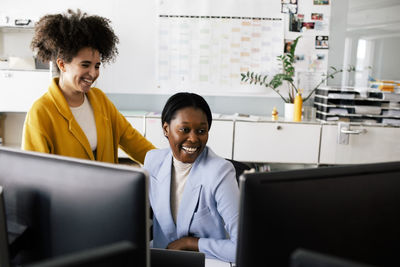 Happy female colleague sitting with businesswoman looking at computers in coworking office