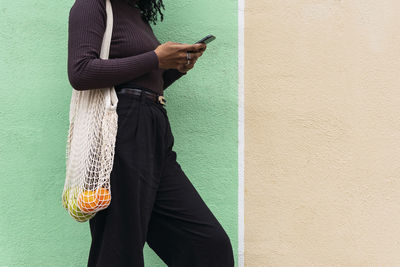 Woman with mesh bag of fruits using smart phone leaning on wall