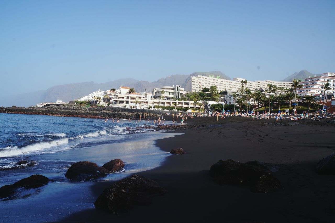 clear sky, water, building exterior, sea, beach, architecture, built structure, blue, copy space, shore, coastline, sand, city, vacations, incidental people, large group of people, scenics, mountain, residential structure