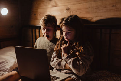 Sibling using laptop while sitting on bed against wall in bedroom