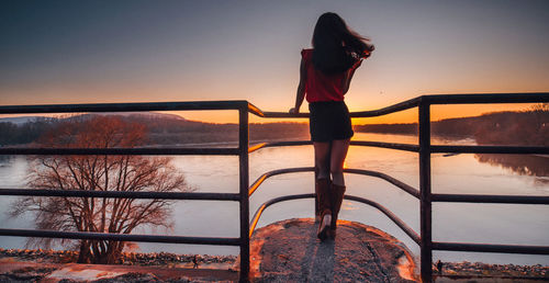 Rear view of man standing by railing against lake during sunset