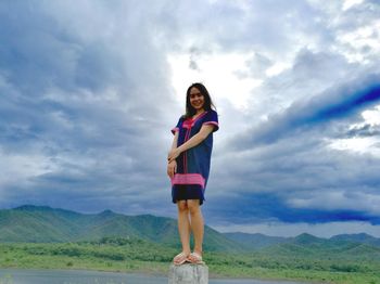 Low angle portrait of smiling young woman standing on bollard against cloudy sky