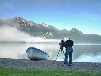 Man hiker is taking photo of ship at mountain lake shore. silhouette at fishing paddle boat at lake